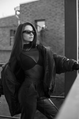 Stylish fashionable woman with makeup and long straight brunette hair in black leather bomber, joggers, high boots, bra, net body and sunglasses standing at red urban metal stairs