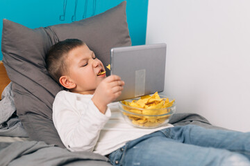 lazy young boy relaxing on bed in his room and using digital tablet and eating junk food