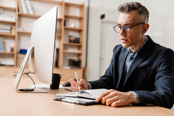 Serious grey man working with computer and planner in office