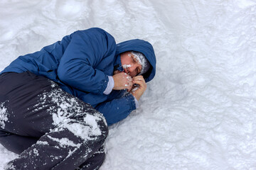 Frozen man in a blue jacket and hat lying down covered snow and frost, trying to stay warm on a...
