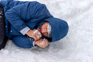 Frozen man in a blue jacket and hat lying down covered snow and frost, trying to stay warm on a...