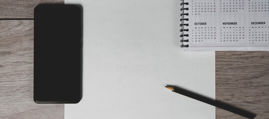 Top view of workplace. Empty paper blank, spiral calendar, black pencil and phone on wooden table.