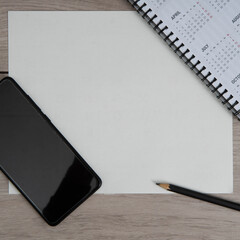 Top view of workplace. Empty paper blank, spiral calendar, black pencil and phone on wooden table.