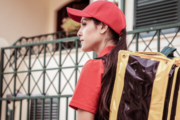 Confident Latin courier delivering order and looking away. Professional young beautiful deliverywoman carrying yellow backpack and wearing red uniform. Food delivery service and post concept