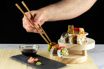 Hand of person holding chopsticks to pick up a sushi roll from a wooden tray with sauce and wasabi.