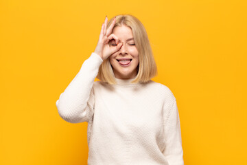 blonde woman smiling happily with funny face, joking and looking through peephole, spying on secrets