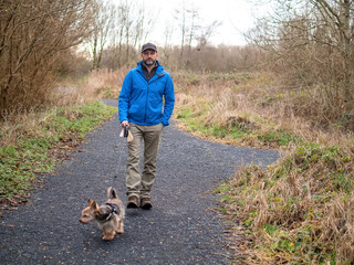 Man and small yorkshire terrier in a park on a small walking path. Animal care and friendship concept. The model in his 40s wears blue jacket and black baseball hat