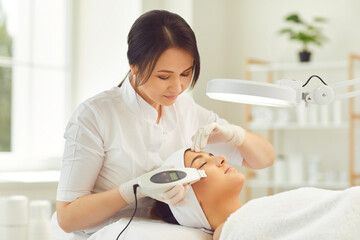 Smiling dermatologist making ultrasound apparatus facial cleaning for young woman