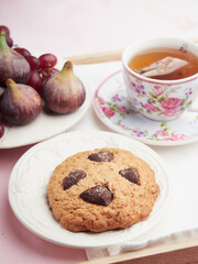 oatmeal biscuit with chocolate chips next to a cup of tea decorated with flowers and fresh fruit on a plate. Figs and grapes.