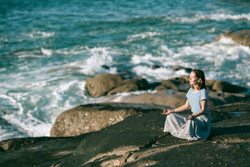 Young yoga woman meditates in the lotus position sitting on rocks on the Atlantic ocean coast.