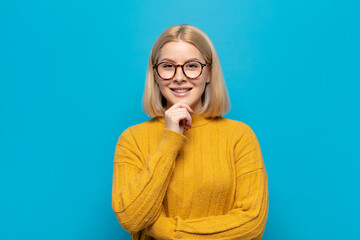 blonde woman looking happy and smiling with hand on chin, wondering or asking a question, comparing...