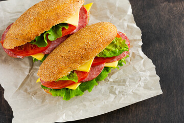 Healthy sandwiches with bran bread, green lettuce, cheese, red tomato and sliced salami on parchment paper and rustic wooden stand. Breakfast concept. Top view.