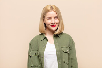 blonde woman looking happy and friendly, smiling and winking an eye at you with a positive attitude