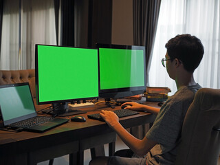 Rear view of an Asian boy wearing glasses working on three computer screens at home. The screens are with green screen.