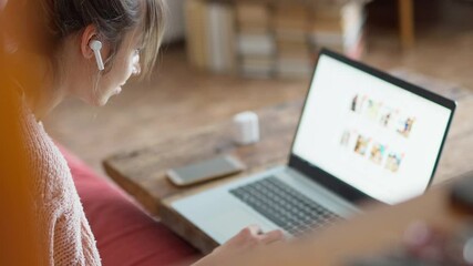 young unrecognizable woman with wireless earphones sits at wooden desk at home, browsing online store on laptop and making online order from home by phone. focus on earphone - Powered by Adobe