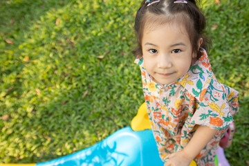 Close up and selective focus with blurred background for top-view shot of adorable asian preschool girl looking with eye contact on slider in grass playground. She shows joyful and positive emotion