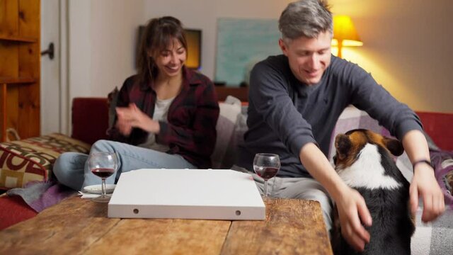 unfocused lovely happy family, man and woman sits on couch in cozy living room at evening with pizza and wine. funny ridiculous welsh corgi dog trying to jumping to couch, man helping him. focus on