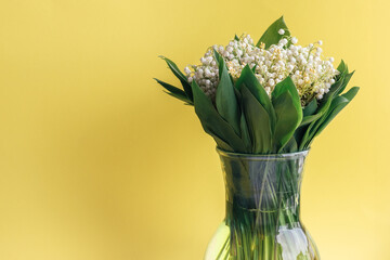 Delicate bouquet of white lilies of the valley in green leaves in a glass vase on a bright yellow background with copy space