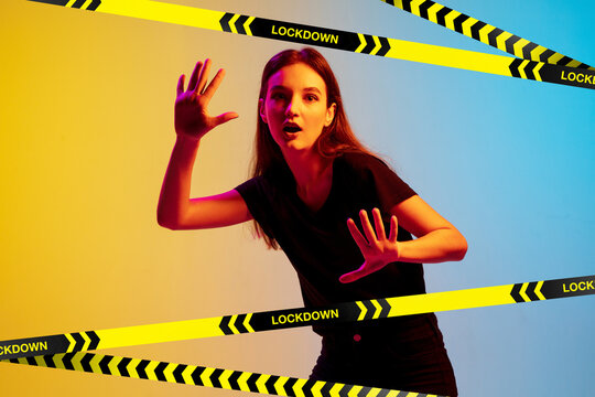 Young Sportive Woman Posing On Gradient Neon Background Behind Limiting Tapes With Word Lockdown. Cancelling Of Gyms, Blocking Social Group Meetings During Covid Pandemic, Isolation Concept.