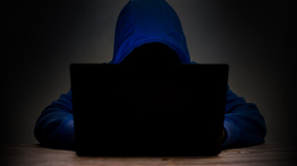 Man wears a blue hooded shirt in front of a laptop computer. Hacking ideas, computer theft to damage