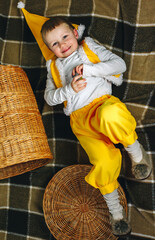 Little boy 4 years old lay on home plaid in yellow gnome elf outfit with cap and straw basket in short pants, turtleneck shirt, tights and fluffy slippers, warm cozy colors