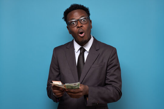 African Man In Suit Holding And Counting Money. Studio Shot On Blue Wall.