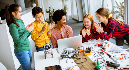 Happy kids with their African American female science teacher with laptop programming electric toys and robots at robotics classroom