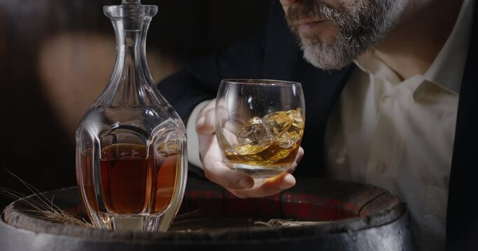 Man in a suit and beard (sommelier) takes a glass of whiskey on ice from a vintage whiskey barrel and shakes the glass to spread the smell of whiskey. Slow motion 4K, 150 fps, Blackmagic Ursa Pro G2. 