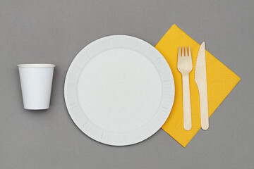 White paper dish, paper cup, wooden fork and knife on orange napkin on gray background, top view. Set of eco friendly disposable tableware from natural material.