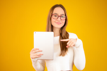Naklejka premium Photo of happy young girl standing isolated over yellow wall background. Looking aside using tablet computer.