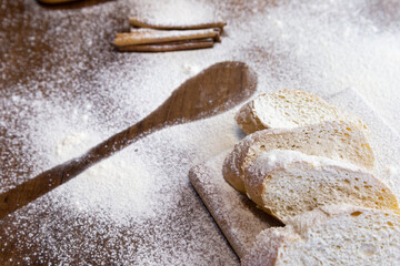 Sliced French baguette on a wooden board, cinnamon sticks and a trail of a large spoon, heavily floured on a wooden table.  