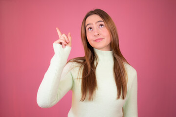 Happy beautiful young woman in green sweater is pointing up, looking away and talking. Waist up studio shot on pink background.
