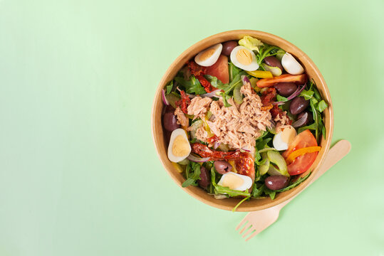 Fish diet salad with tuna, tomatoes, arugula on green background with wooden fork. Healthy lunch. Concept environment protection, plastic free. Food delivery in disposable plate of craft paper.