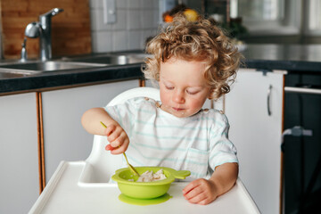 Cute adorable Caucasian curly kid boy sitting in high chair eating cereal puree with spoon. Healthy eating for kids children. Toddler eating independently. Candid real authentic moments.