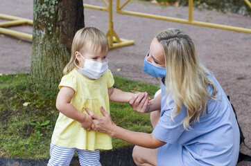 Fototapeta premium Woman and child girl in the park wearing medical face mask. Mother and her little daughter in surgical mask, closed park for visitors. Wearing surgical masks in public. 