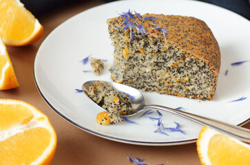 Poppy seed and orange peel vegan egg free and dairy free cake, bite on the orange plate with blue linen textile and poppy head nearby, closeup, copy space, vegan food and pastry concept