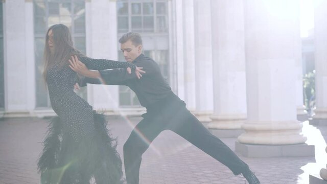 Handsome young man dancing paso doble with partner in sunny city at urban building with columns. Confident professional Caucasian ballet dancers performing latin ballroom dance outdoors.