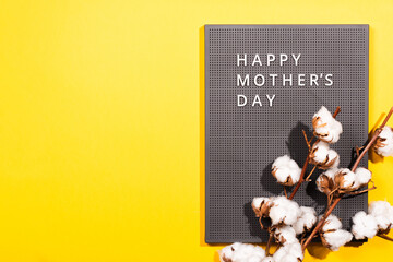 Grey plastic letter board with white quotes Happy Mother's Day, and cotton branch on a illuminating yellow background.