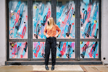Girl wearing COVID-19 mask is posing full length by graffiti in Manhattan, New York. She is wearing...