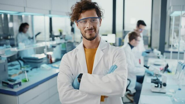 Medical Science Laboratory: Handsome Young Latin Scientist Wearing White Coat Puts on Safety Glasses, Smiles Looking at Camera. Young Bio Technology Research Specialist. Medium Closeup Portrait Shot