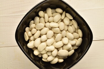 Dried grains of organic white beans in a black ceramic bowl on a wooden table, top view.