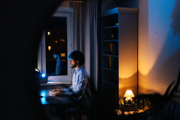 Remote view of young freelancer working on computer and typing text on wireless keyboard while sitting at desk at home office workplace at late night in dark room. Concept of remote working.