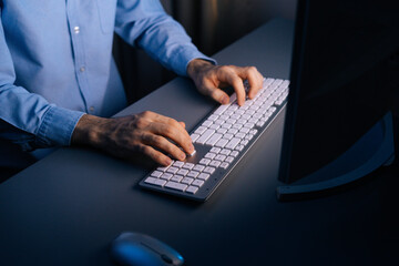 Top view of hands of unrecognizable man using computer typing message on wireless keyboard at dark room. Close-up view of working on pc. On monitor screen change of images. Concept of remote working.