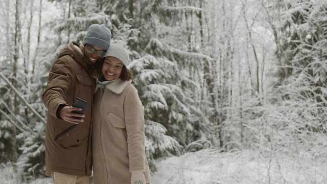 Medium shot of young multiethnic man and woman standing in winter forest, hugging and making photos using smartphone