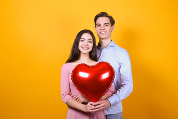 Fototapeta na wymiar Happy Valentine's day concept. Studio shot of couple in love holding a heart shaped balloon, showing affection. 14th february - the lovers day. Yellow wall background, copy space, front view portrait.
