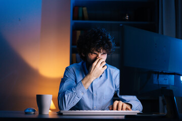 Close-up view of disappointed upset young man touching his head with face like everything failed while sitting at desk with computer at home. Displeased bearded curly guy showing facepalm gesture.