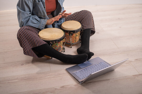 A faceless woman sits on the floor at home and watches educational videos of playing mini bongs. Girl remotely learns to play traditional ethnic drums.