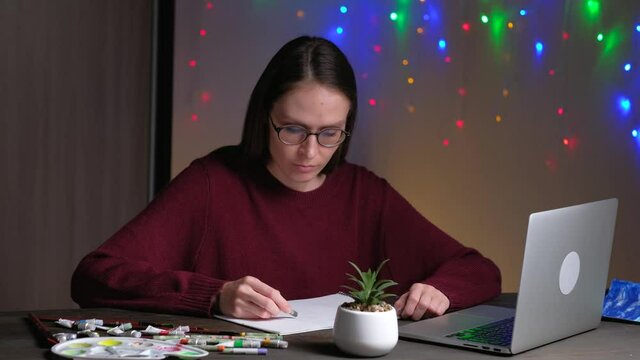 Young female learn draw remotely using laptop and colored pencil, sitting on the table in evening at home on background of christmas decorations garlands. Concept completing a task at home online.