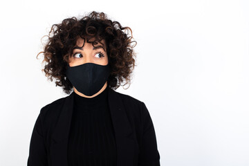 young beautiful caucasian woman wearing medical mask standing against white wall with thoughtful expression, looks away keeps hands down bitting his lip thinks about something pleasant.