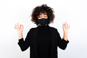 young beautiful caucasian woman wearing medical mask standing against white wall doing yoga,...